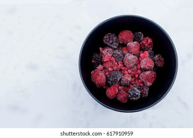 Frozen Berries in a black bowl on a marble surface (strawberries, blackberries, raspberries)