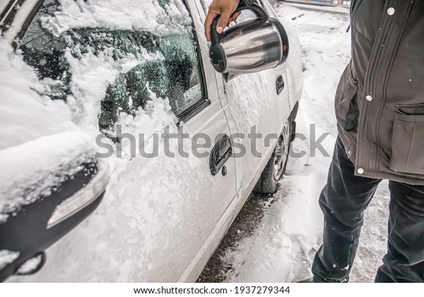 A\
frozen car in the snow in a parking lot in a city. A man pours hot\
water from a kettle on a frozen door and a car\
lock.
