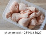 Frozen chicken drumsticks in a plastic container defrosting. On a wooden chopping board