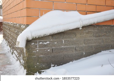 A Frozen Downspout Rain Gutter Pipe With Snow And Icicles Along The House Foundation, Basement, House Wall In Winter.