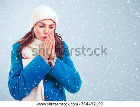 Frozen girl in blue coat, white hat and scarf heats hands among winter snowstorm. 