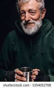 Frozen homeless bearded old man with grey hair and wrinkled face, looks at camera with grateful expression and holds a steel mug with coins given as alms, posing at studio over black background