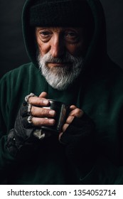 Frozen homeless bearded old man with grey hair and wrinkled face, looks at camera with grateful expression and holds a steel mug with coins given as alms, posing at studio over black background