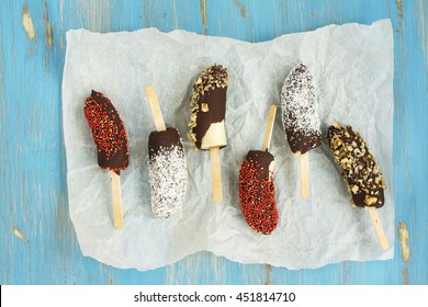 Frozen Homemade Banana Pops Covered With Chocolate, Sprinkles, Nuts And Coconut Chips Over Blue Wooden Table. Top View