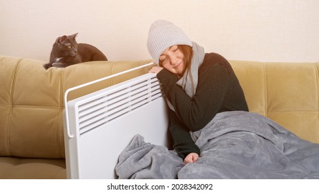 Frozen Lady With Grey Plaid In Scarf And Knitted Hat Leans On Heater Sitting Near Cat On Soft Modern Sofa In Living Room.