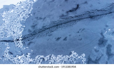 Frozen Lake Pictured From Above With Drone. Top View Of Melting And Cracked Ice On Dark Water With Boat Traces And Spots In Winter As An Abstract Background. Uneven Surface.
