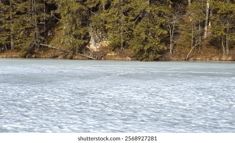 A frozen lake stretches towards a dense, evergreen forest. Snow covers the ice, creating a rippling texture. Lacul Rosu - Romania - Powered by Shutterstock