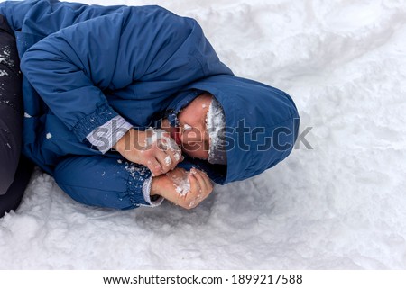 Frozen man in a blue jacket and hat lying down covered snow and frost, trying to stay warm on a very cold winter day, snow falls around him. Sick mountaineer with hypothermia on snow during the day.