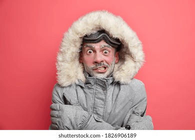 Frozen man trembles from cold has red face covered by ice frosted beard wears jacket with hood needs to warm during winter expedition poses over coral background. Cold weather low temperature - Powered by Shutterstock