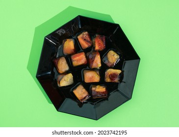 Frozen Mixed Fruit Cubes Lie On A Black Ceramic Plate.. Summer Refreshment Concept, Flat Lay Composition On Bright Pastel Green Background