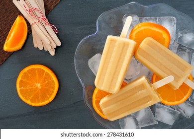 Frozen orange yogurt popsicles in an ice filled bowl with fresh fruit slices against a slate background