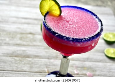 Frozen prickly pear margarita in a salted, blue rimmed glass with lime slice on a rustic wooden surface