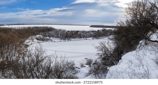 Frozen quarry surrounded by leafless trees and snowy hills under a cloudy winter sky, creating a calm and expansive cold-season landscape