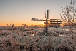 Frozen Reeds And Grass Field With An Old Polder Mill During Sunrise. The Sun Shines Through One Of The Blades. Kalenberg, Overijssel In The Netherlands.