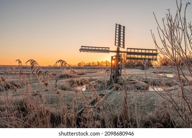 Frozen reeds and grass field with an old polder mill during sunrise. The sun shines through one of the blades. Kalenberg, overijssel in the Netherlands.