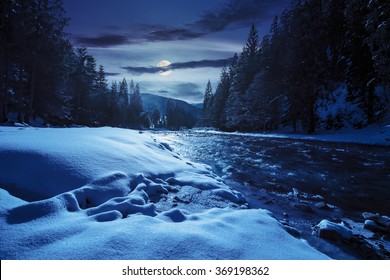 Frozen River Among Conifer Forest With Snow On The Ground In Carpathian Mountains At Night In Full Moon Light