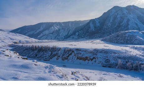 A frozen river winds in a valley between steep banks. Fluffy hoarfrost on dry grass and on the ground. A picturesque mountain range against the blue sky. Altai - Shutterstock ID 2220378939