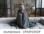 Frozen. Sad latina female sit on couch at freezing cooled studio flat in warm cap and blanket shiver tremble with cold. Unhappy young lady spend time at home feel bad suffer of heating system problems