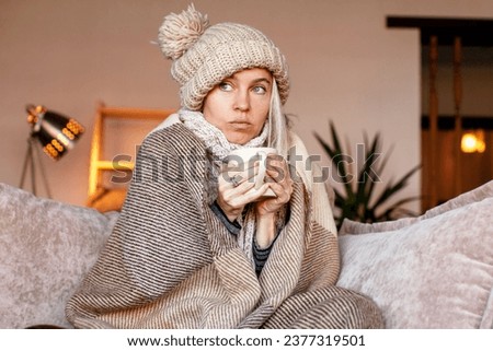 Frozen. Sad woman sit on couch at freezing cooled house in warm cap and blanket shiver tremble with cold. Unhappy middle aged lady spend time at home feel bad suffer of heating system problems