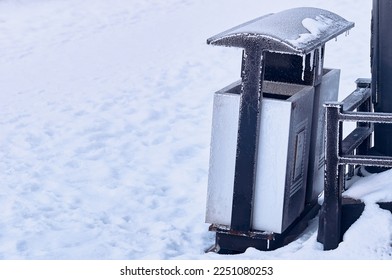 Frozen trash can on pure white snow. Icicles and frost on black metal. Climate change concept. Winter chill. Waste management in the urban environment. Strong cooling. Close up.