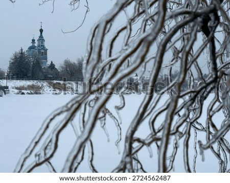 Frozen tree branches covered with ice in the foreground, winter landscape with a church in the background across a frozen river.