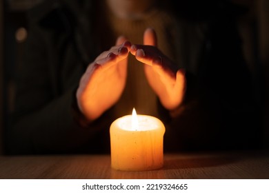 frozen woman in warm jacket tries to heat palms over burning candle sitting in cold room without central heating. No heating - Shutterstock ID 2219324665
