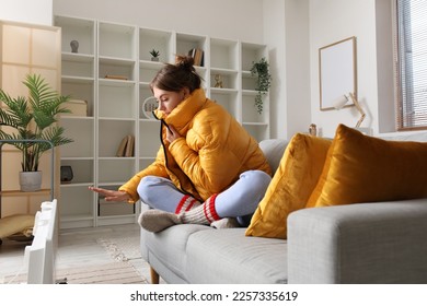 Frozen young woman in down jacket warming near radiator at home - Shutterstock ID 2257335619