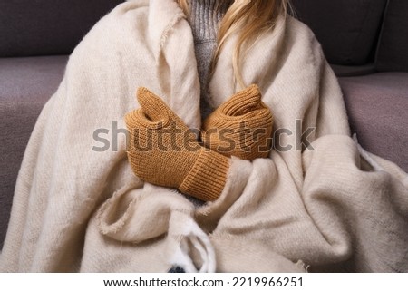 Frozen. A young woman sits on the floor in an apartment in a warm sweater, plaid and mittens and is shivering from the cold. 
