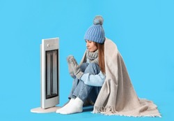 Frozen Young Woman Warming Near Electric Heater On Blue Background