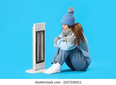 Frozen young woman warming near electric heater on blue background - Shutterstock ID 2257335611
