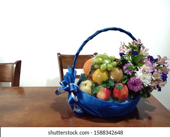 A fruit basket contains apple, grapes, kiwi, oranges, pear in a wrapped blue ribbon basket with flower on the wooden table and a white wall. - Shutterstock ID 1588342648
