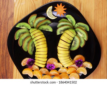 Fruit dessert made from sliced bananas, kiwi, tangerines, grapes laid out on a plate in the form of palms and tropical trees.