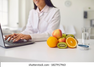 Fruit, a glass of water and a tape measure lie on a white table in a bright dietitian's office against the background of a nutritionist who writes a diet plan. Female doctor using laptop for her work.