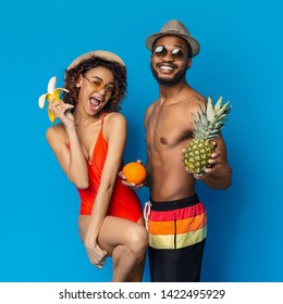 Fruit nutrition. Happy african girl and guy enjoying banana, orange and pineapple, blue studio background