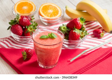 Fruit Smoothie Glass On Red And White Stripes Napkins Around Spread Ingredients: Strawberries, Sliced Orange, Bananas, Blurred On White Wood Background. Strawberry Orange Banana Smoothie. Horizontal. 