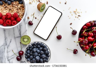 Fruits and berries breakfast delivery composition blueberry  raspberry cherries kiwi and mobile cellphone with blank screen for text copy space top view on white background