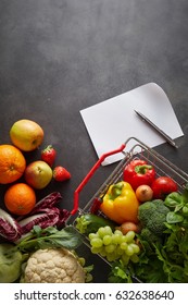 Fruits and vegetables in shopping basket and on table, near blank white shopping list and pen and copy space over clean blackboard background
