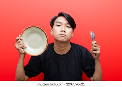 Frustated Asian Man With Sad Expression Holding Empty Plate, Spoon And Fork. Fasting And Diet Concept. Man Is Starving There Is No Foods.