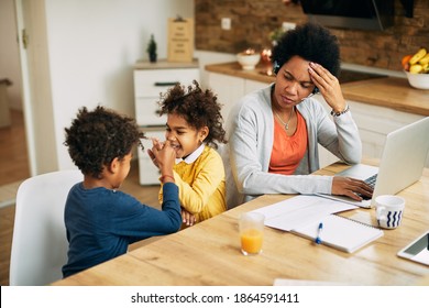 Frustrated African American Mother Working On A Computer While Her Kids Are Playing And Making Noise At Home. 