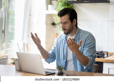 Frustrated Angry Young Man In Eyeglasses Looking At Laptop Screen, Feeling Nervous About Bad Device Work Or Poor Internet Connection In Kitchen. Unhappy Shocked Guy Reading Message With Bad News.