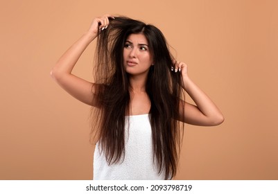 Frustrated Armenian Woman Showing Her Damaged Long Locks On Beige Studio Background. Young Lady Having Bad Hair Day, Upset Over Her Messy Hairdo. Hairdressing Services Concept