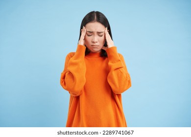 Frustrated asian woman holds hands on head, has headache, looks miserable, cant handle pressure, feels distressed, stands over blue background. Mental health concept.