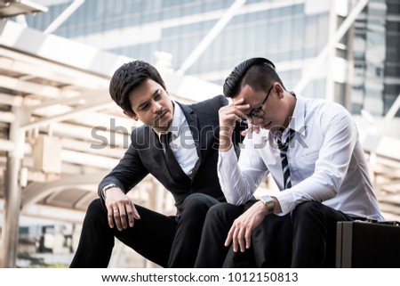 Frustrated Asian young business man sitting outdoor office after he had failed in new project, his colleagues tried to reassure his.