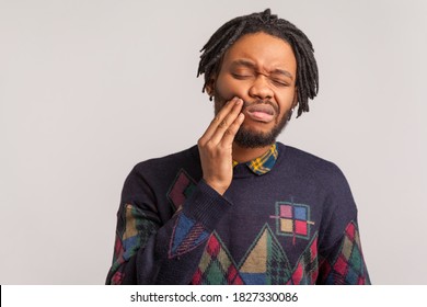 Frustrated Bearded African Man With Dreadlocks Touching Cheek And Wincing In Pain Feeling Terrible Tooth Ache, Gingivitis, Periodontal Disease. Indoor Studio Shot Isolated On Gray Background