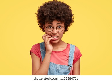 Frustrated beautiful African American female feels anxious and surprised, bites finger nails in puzzlement, looks nervously at camera, worries before presenting project work in front of audience