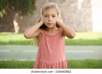 Frustrated Beautiful Caucasian Little Kid Girl Wearing Dress Standing Outdoors Plugging Ears With Hands Does Not Wanting To Listen Hard Rock, Noise Or Loud Music.