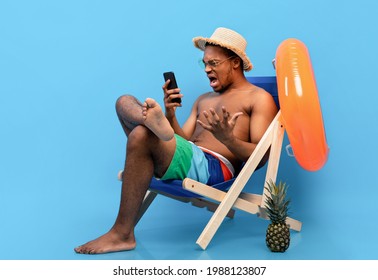 Frustrated Black Guy Shouting At Smartphone, Feeling Stressed, Having Bad Internet Connection On Summer Vacation, Sitting In Lounge Chair With Mobile Device, Blue Studio Background