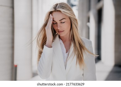 Frustrated blonde caucasian young woman walking eyes closed with upset facial expression touching head, depressed, having financial problems. Beautiful swedish woman having headache outdoors.