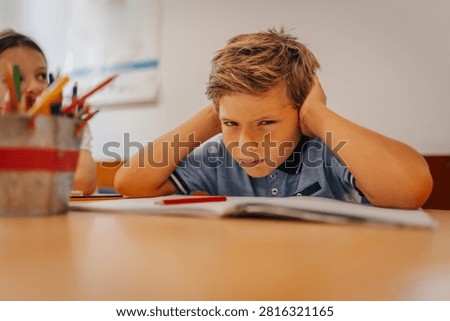 Frustrated boy student sitting at elementary school desk, covering ears, experiencing stress and a learning difficulty in class
