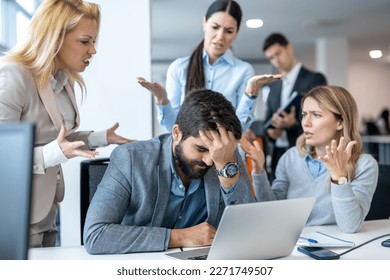 Frustrated businessman covering face with hand, having serious problems while his co-workers making him feels guilty for not doing his job properly at office. Business conflict and overwork concept.